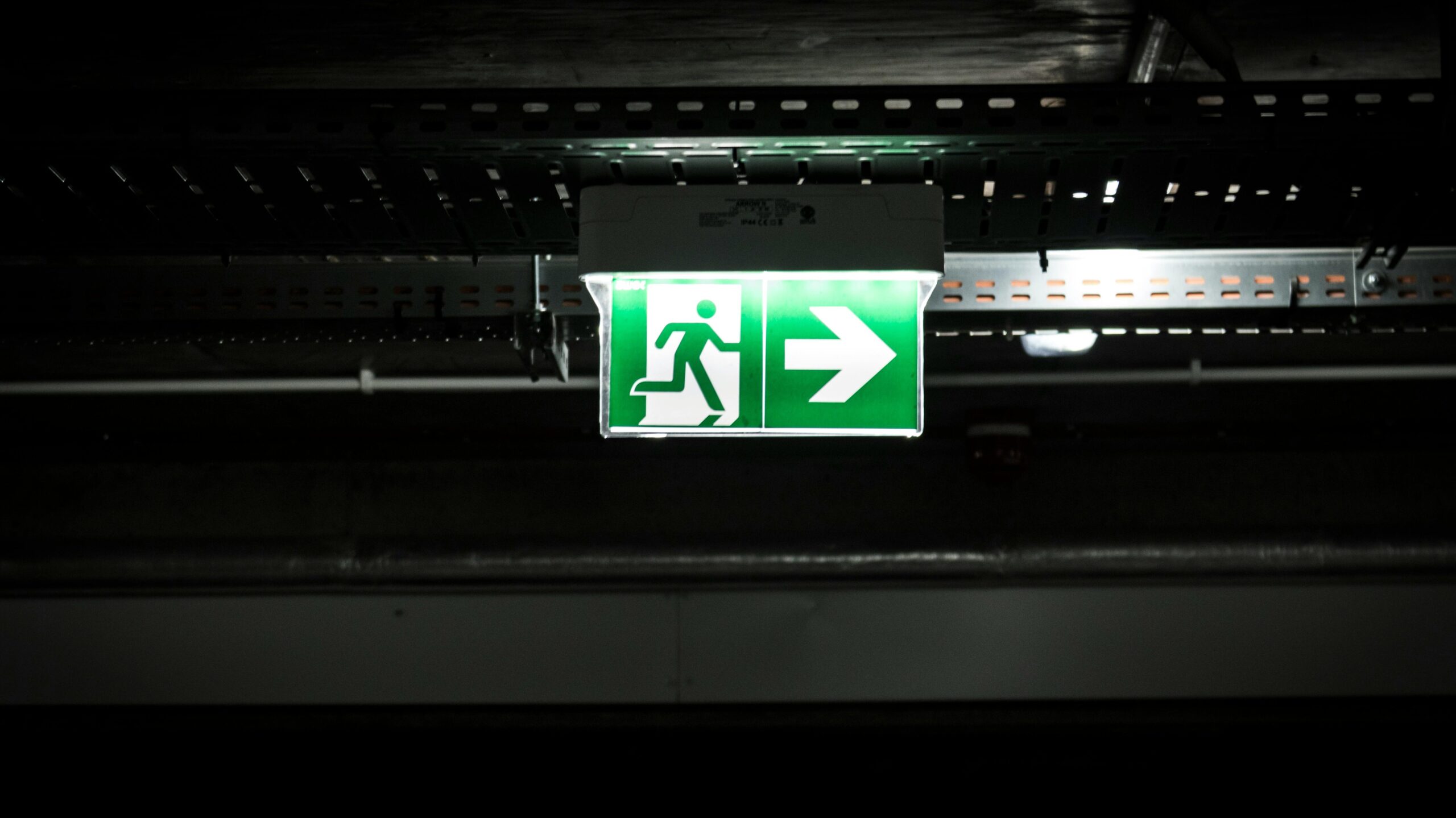 Bright green emergency exit sign with arrow in a dark industrial setting, indicating evacuation route.